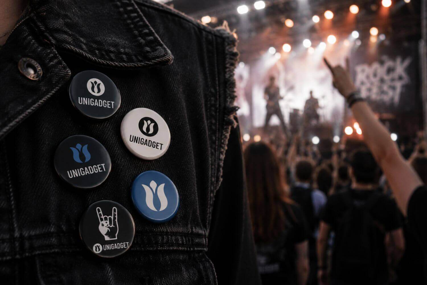 Dettaglio di un giubbotto di jeans nero decorato con diverse spille personalizzate rotonde con il logo Unigadget e il simbolo delle corna rock, con il palco sfocato sullo sfondo.
