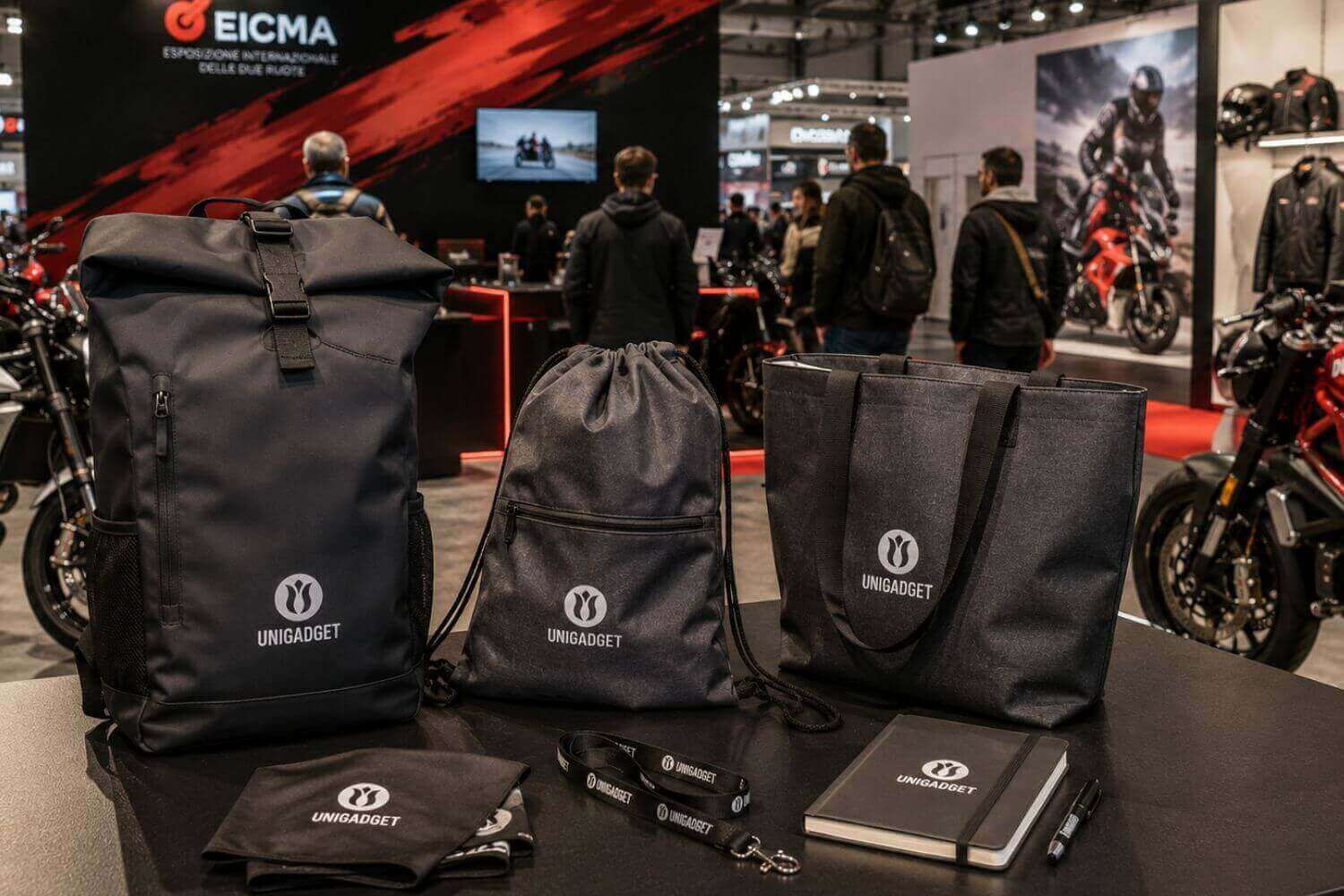 Zaino roll-top, sacca a zainetto e shopper personalizzati con il logo Unigadget, esposti allo stand della fiera EICMA di Milano insieme a un taccuino, una penna e un lanyard, con motociclette in mostra sullo sfondo.